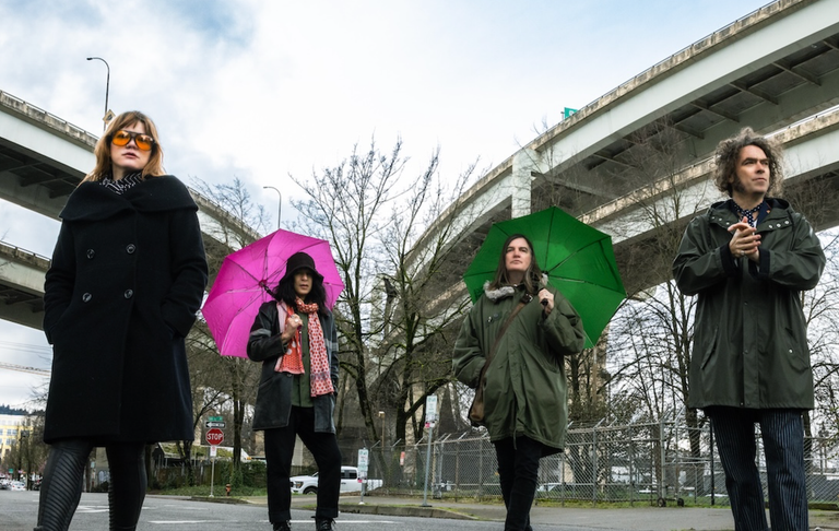 American rock band The Dandy Warhols standing outdoors near elevated overpasses with pink and green umbrellas in a press photo.